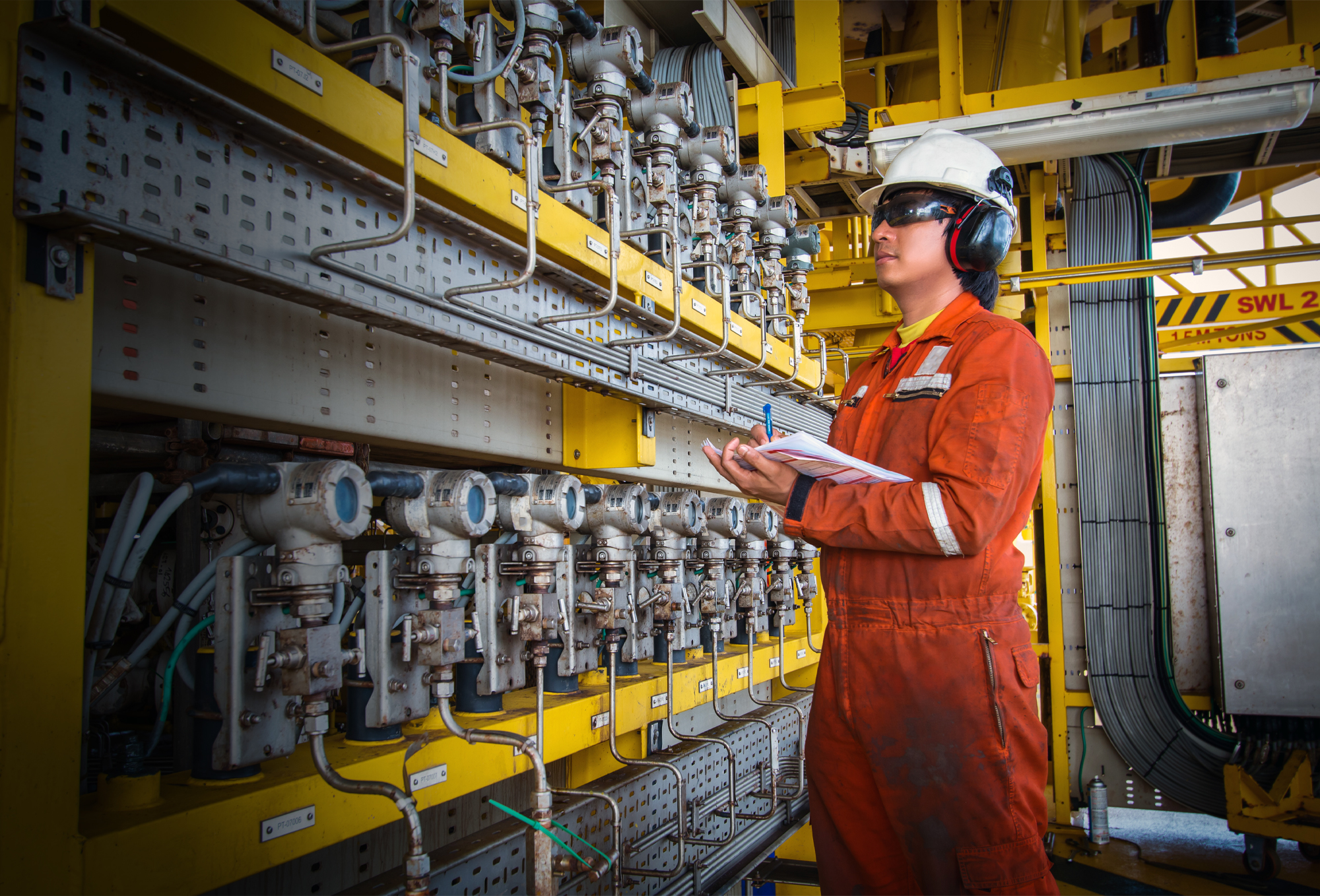 Technician in PPE Standing with Clipboard in Front of Rows of Pressure Gauges